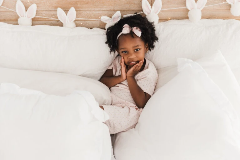 Young girl in pajamas sits in a white bed with bunny-ear decorations in the background, looking directly at the camera.