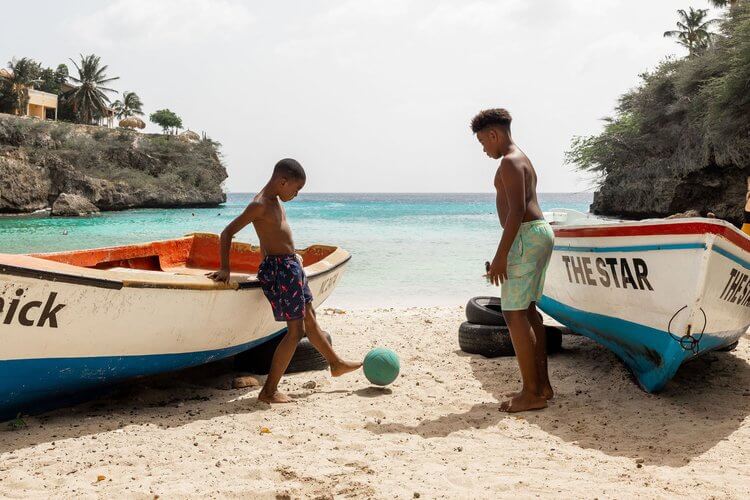 Two boys play with a ball on a sandy beach near two beached boats, with a backdrop of turquoise water and lush coastal foliage.