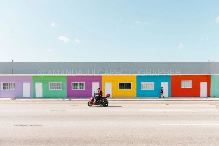 Man rides red scooter past colorful buildings under bright blue sky on city street, with woman walking in the background.