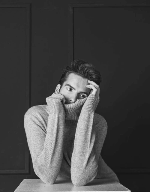 Black and white portrait of an adult male partially obscured by a gray turtleneck sweater against a dark gray paneled background.