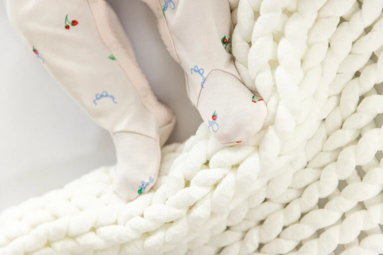 Baby's legs and feet clad in patterned pajamas rest on a chunky knit blanket, shot in bright, soft focus to highlight texture.