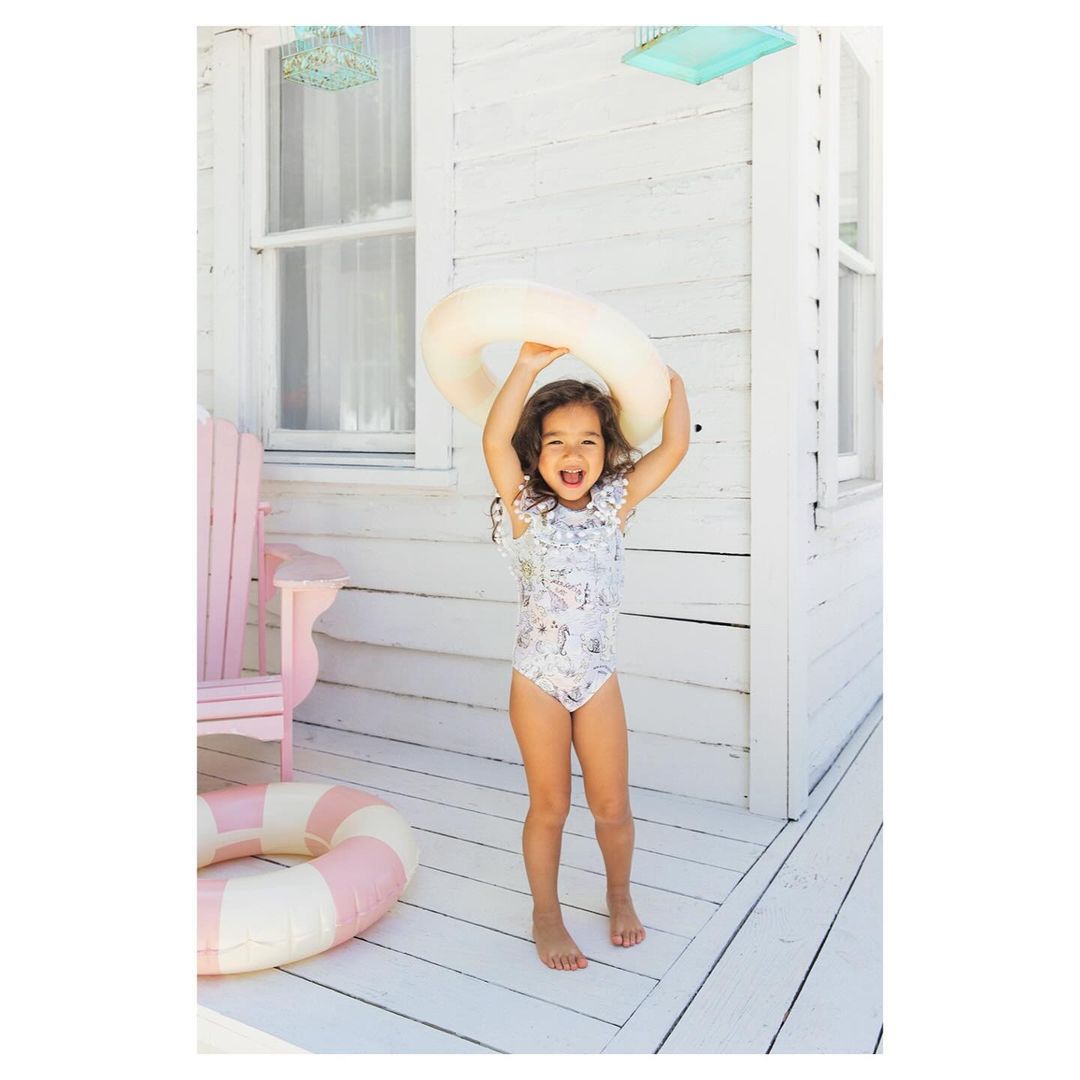 Smiling young girl in a patterned swimsuit stands on a porch holding a white swim ring above her head in a summer scene.
