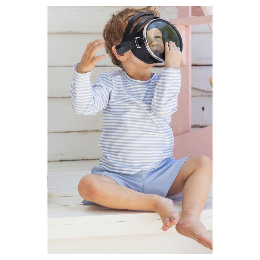 Young child sits outdoors wearing a striped shirt, light blue shorts, and vintage diving mask. Summery scene, light and airy.