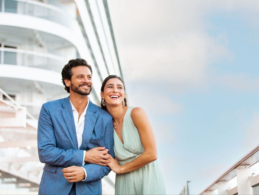 A couple in formal wear stands on the deck of a cruise ship, smiling and enjoying the ocean view. The woman rests her head on the man's shoulder.