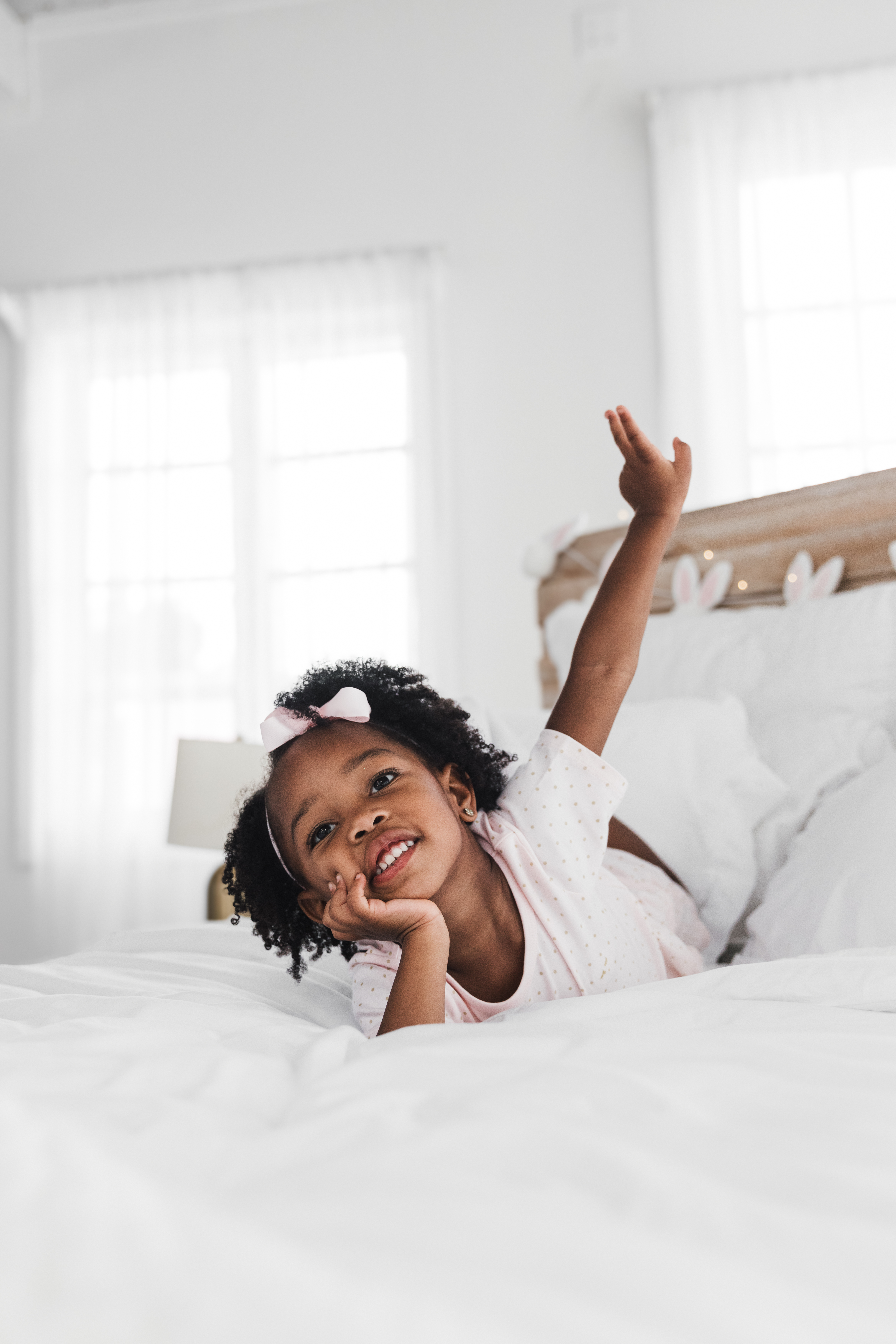A young African-American girl lies on a white bed, smiling with her hand under her chin. A wooden headboard is in the background.