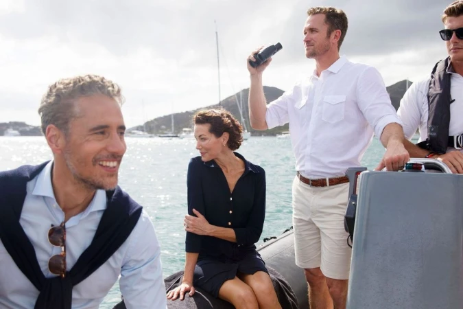 Group of four people on a boat, one using binoculars, water and islands in the background under a cloudy sky. Sailing activity.