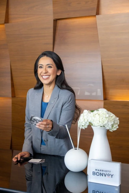 Smiling woman in business suit stands at hotel reception holding loyalty card, with white flower arrangement and wood-paneled wall.