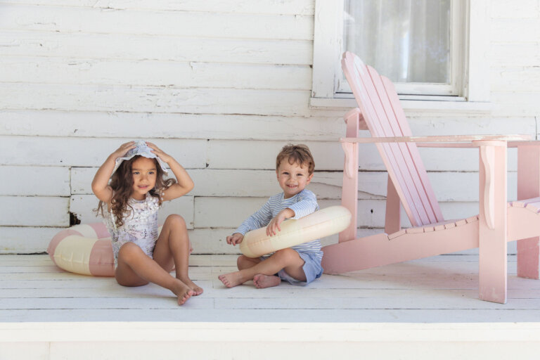 Two children, a girl and a boy, sit on a porch near a pink Adirondack chair with pink and yellow pool floaties and smile at the camera.