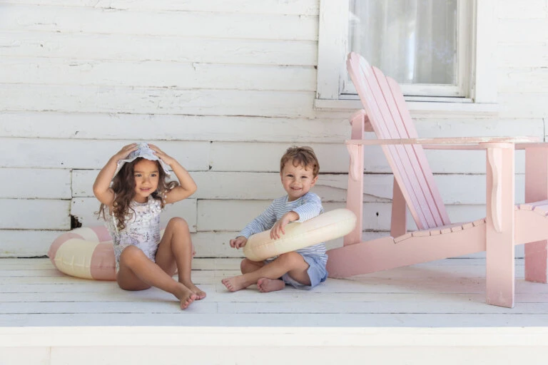 Two children, a girl and a boy, sit on a porch near a pink Adirondack chair with pink and yellow pool floaties and smile at the camera.