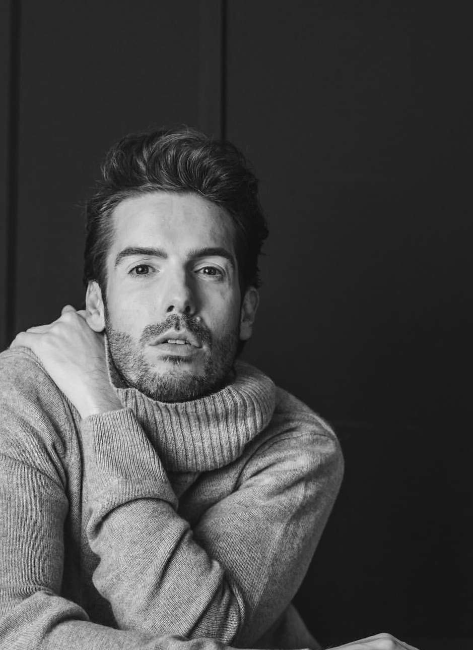 Black and white portrait of a bearded man with dark hair wearing a gray turtleneck sweater, posing against a dark gray background.