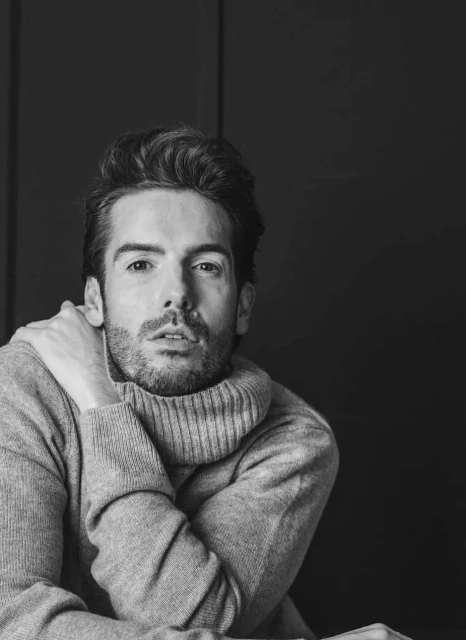 Black and white portrait of a bearded man with dark hair wearing a gray turtleneck sweater, posing against a dark gray background.
