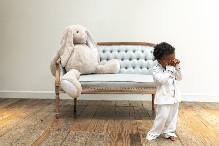 Child in pajamas covering face next to a stuffed bunny rabbit sitting on a tufted loveseat in front of a white wall.