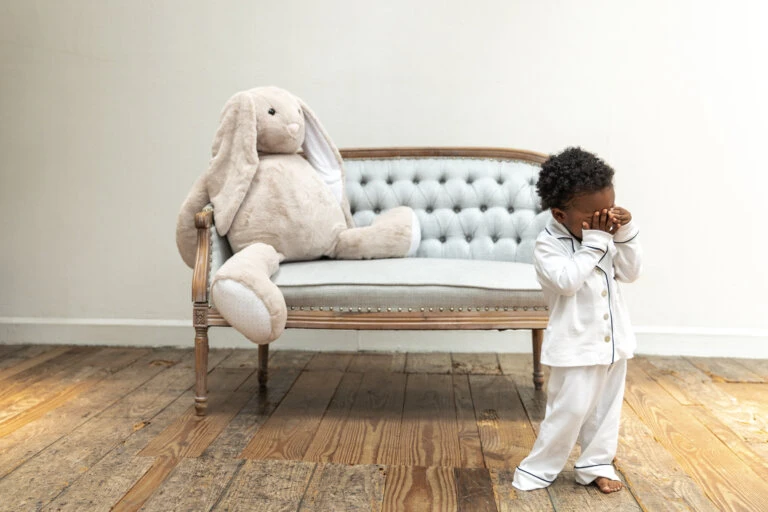 Child in pajamas covering face next to a stuffed bunny rabbit sitting on a tufted loveseat in front of a white wall.