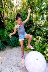 Young child balancing on a pink and white inflatable ball in front of lush tropical foliage.
