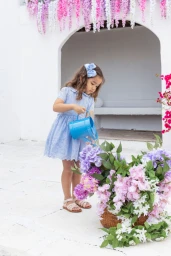 Young child in a light blue dress watering flowers with a blue watering can in front of a white archway.