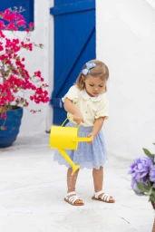 Young child holding a yellow watering can against a white wall with blue doors and pink flowers