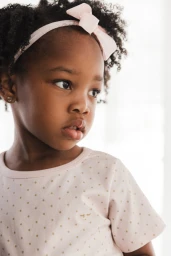 Young child with curly hair wearing a pink headband with a bow and a dotted shirt.