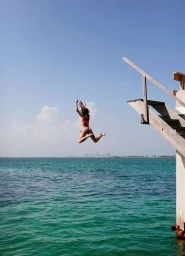 Woman mid-air diving from a wooden platform into turquoise ocean water with a distant city skyline visible on the horizon.