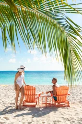 A woman in a white swimsuit and sun hat stands beside a man seated in an orange beach chair on a sandy shore beneath palm fronds.
