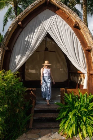 Woman wearing a sun hat and patterned maxi skirt standing in the arched wooden entrance of a thatched bungalow.