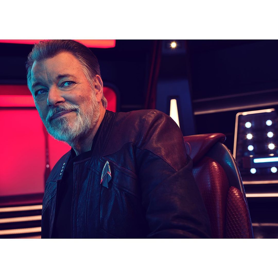 Headshot of Jonathan Frakes wearing a Starfleet jacket, seated in a red chair against a blurred background with blue and red lighting.