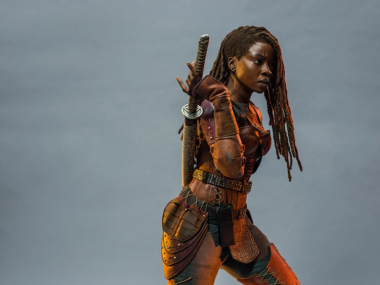 Portrait of a woman with dreadlocks in leather armor with a sword on her back against a grey background in a studio.