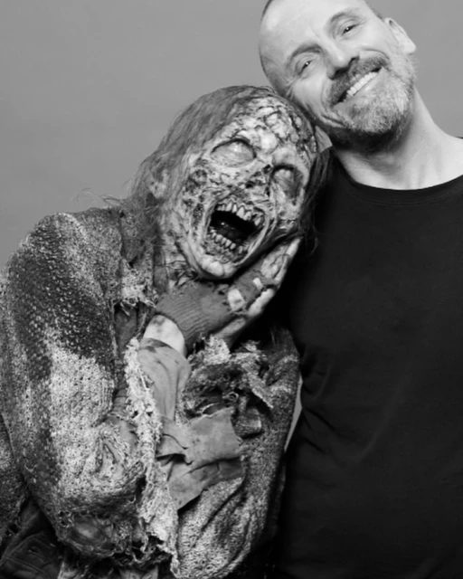 A man in a black shirt smiles while posing next to a zombie with decaying skin and an open mouth in a grayscale studio shot.