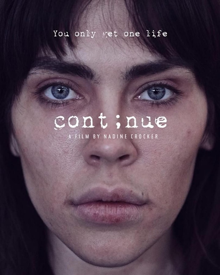 Close-up portrait of a young adult woman with brown hair and striking blue eyes, against a dark background, reminiscent of a film poster.
