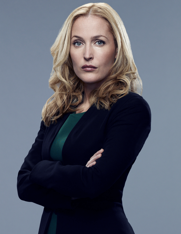 Studio portrait of a blonde woman with blue eyes wearing a dark blazer and green top, arms crossed in front of a grey background.