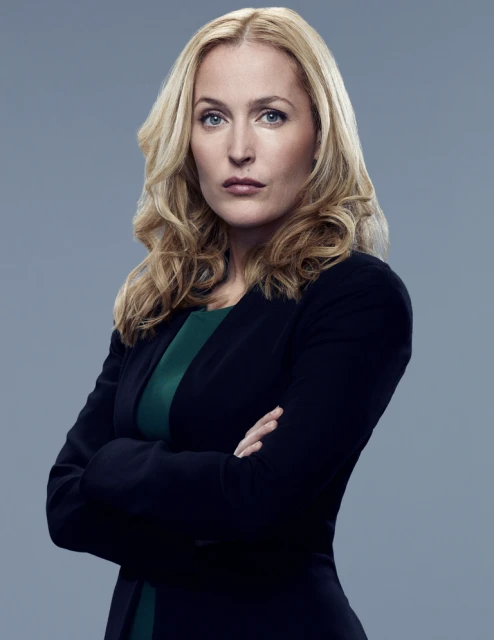 Studio portrait of a blonde woman with blue eyes wearing a dark blazer and green top, arms crossed in front of a grey background.