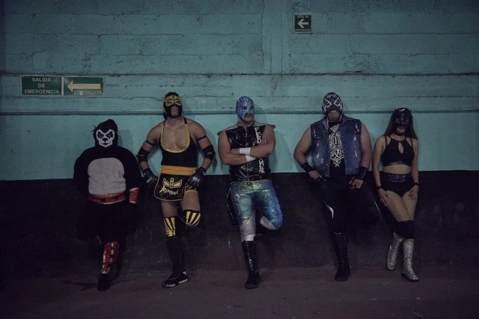 Group of five masked Mexican wrestlers posing against a blue wall, showcasing colorful costumes and theatrical masks in indoor setting.