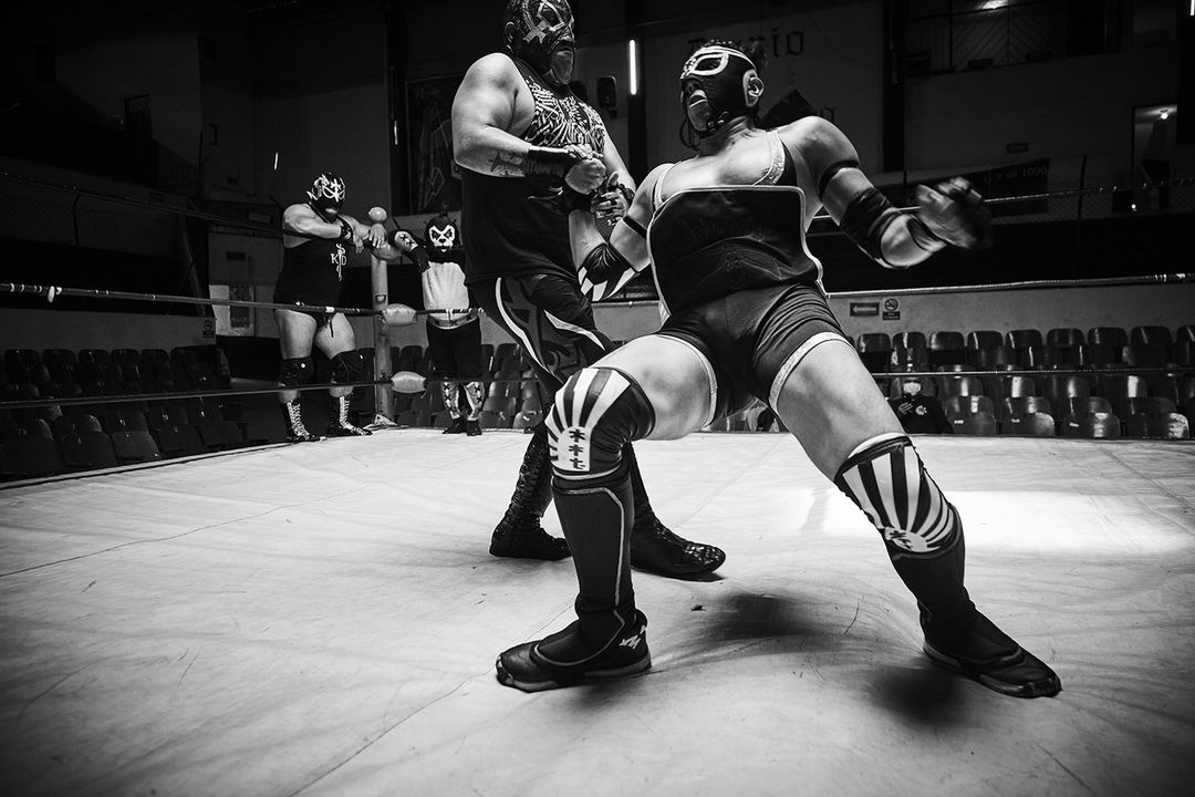 Two masked wrestlers face off in a black and white photo inside of a wrestling ring. Several spectators are in the background.
