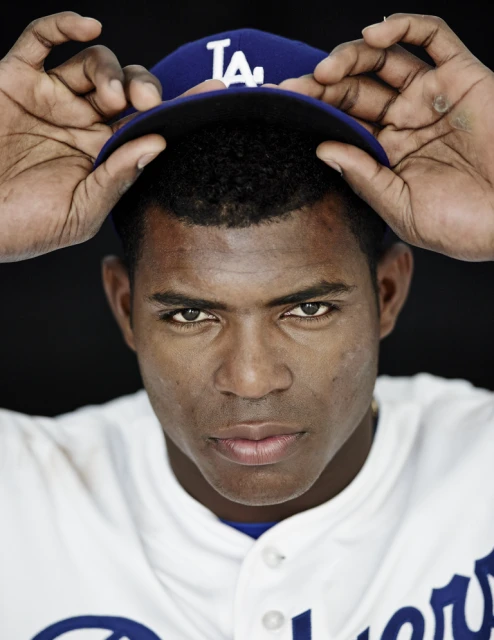 Close-up of an adult male wearing a baseball uniform and adjusting a blue baseball cap with the letter 'LA' on it.