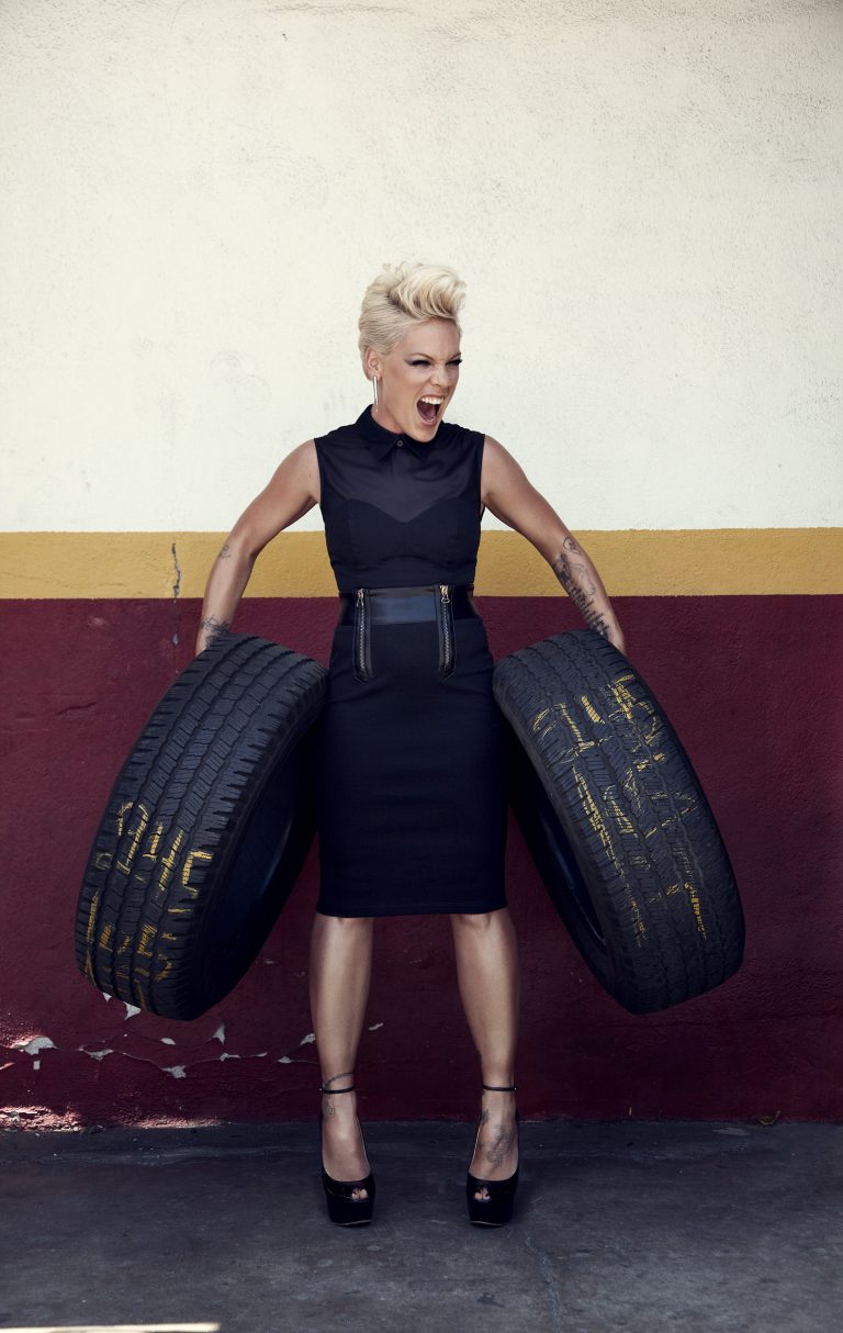 Woman with pink hair and black clothing confidently holds two tires against a cream-colored wall with a horizontal red stripe.