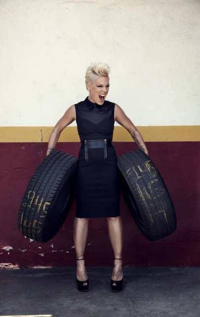 Woman with pink hair and black clothing confidently holds two tires against a cream-colored wall with a horizontal red stripe.