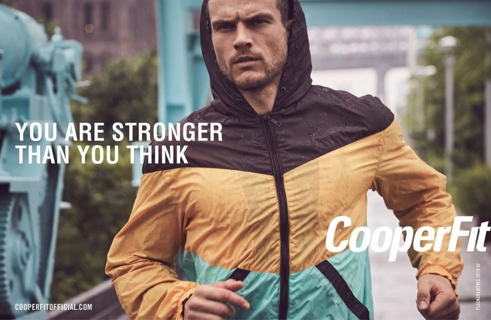 Athletic adult male wearing a hooded windbreaker jacket runs outdoors in the rain, with an urban bridge structure in the background.