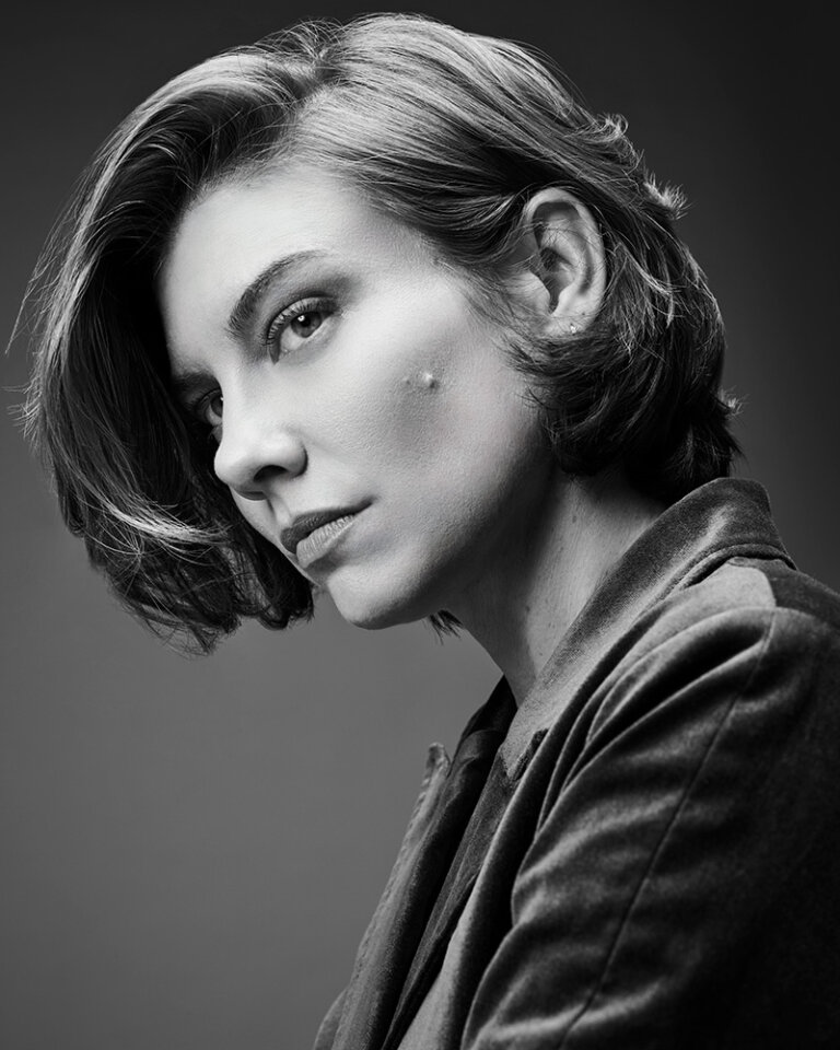 Black and white portrait of a woman with short hair wearing a dark jacket, gazing off to the side in a studio setting.