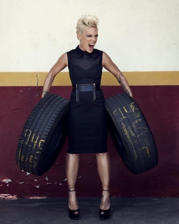 Singer Pink poses with tires in a garage. She is wearing a fitted black dress and platform heels and has a distressed expression.