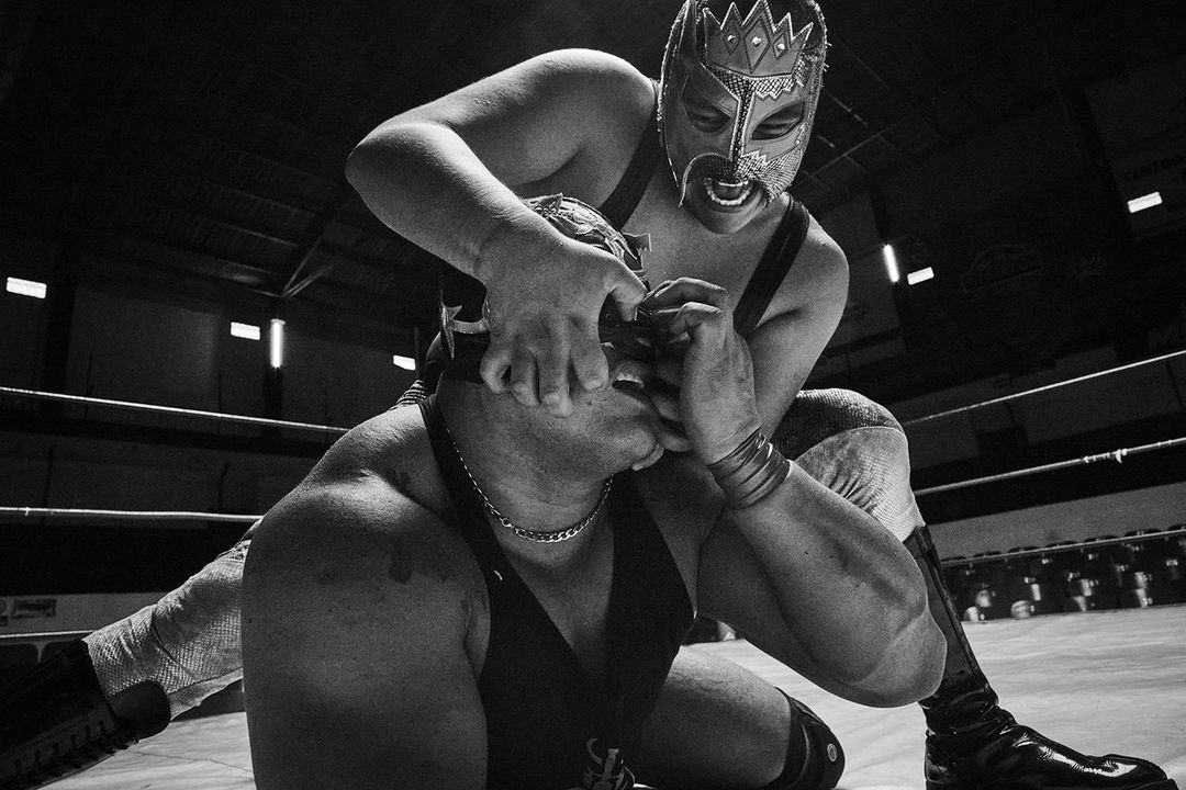 Two masked luchadores wrestling in a ring, one pinning the other by the face, captured in a high-contrast black and white photograph.