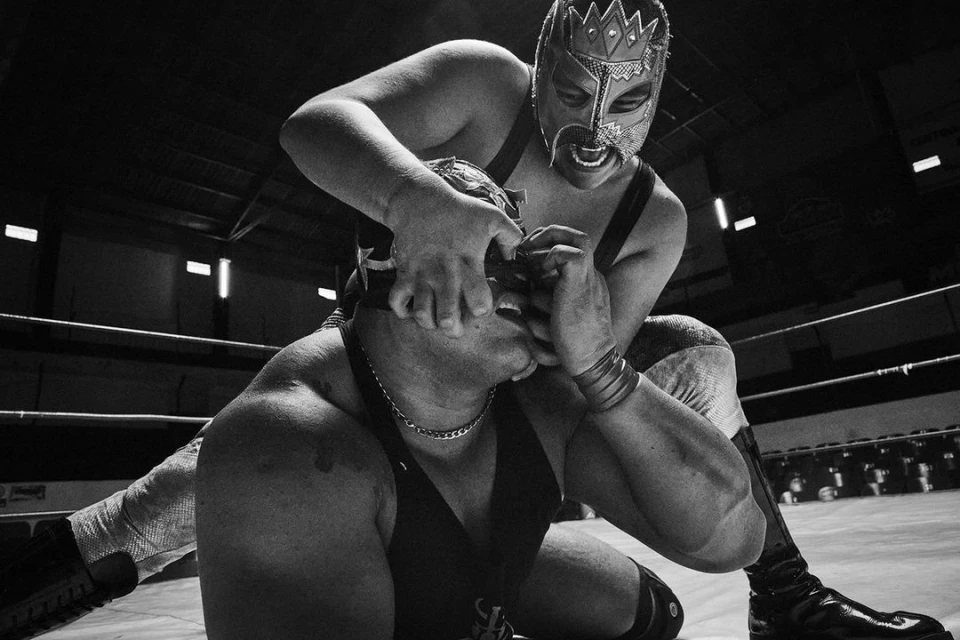 Two masked luchadores wrestling in a ring, one pinning the other by the face, captured in a high-contrast black and white photograph.