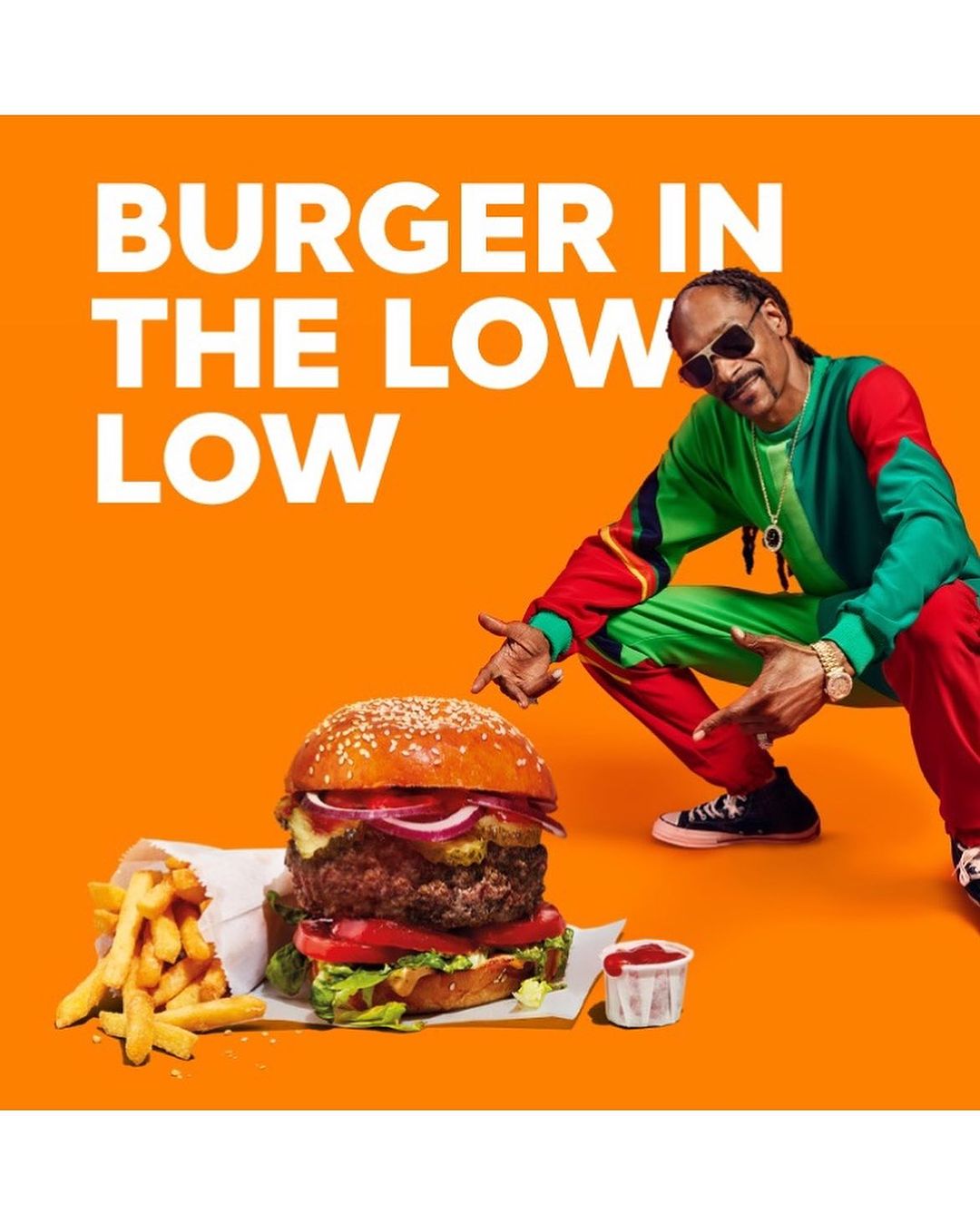 Snoop Dogg squats next to a large burger with fries and ketchup against a bright orange background in a promotional image.