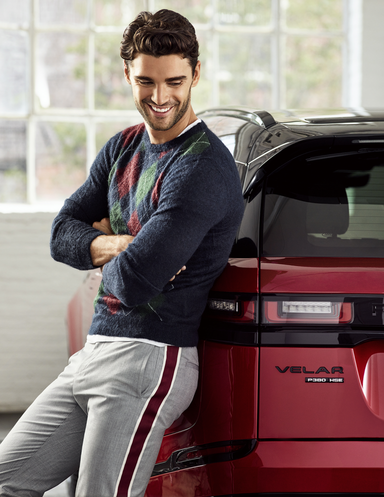 Attractive adult male smiles while leaning against a red Velar P300 HSE car in a brightly lit indoor setting with large windows.