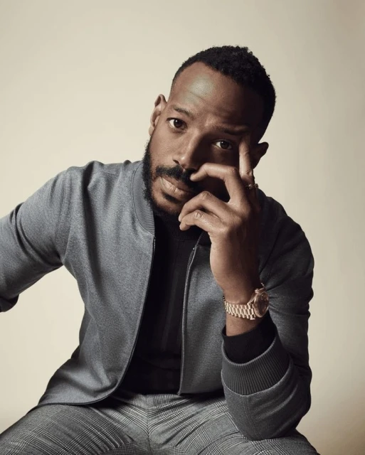 Close-up portrait of a man with a beard wearing a grey bomber jacket and grey plaid pants in a photo studio setting.