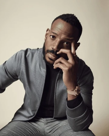 Close-up portrait of a man with a beard wearing a grey bomber jacket and grey plaid pants in a photo studio setting.