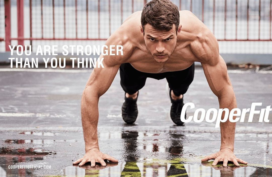Muscular man in athletic wear is doing a push-up outdoors on a wet surface with arms extended, showcasing fitness and upper body strength.