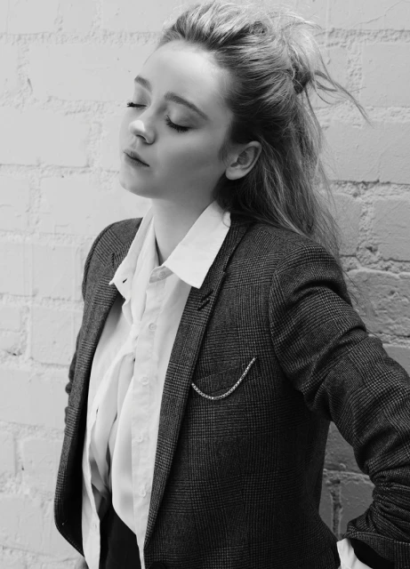 Black-and-white portrait of a young woman in a blazer, eyes closed, leaning against a white brick wall, with soft natural lighting.