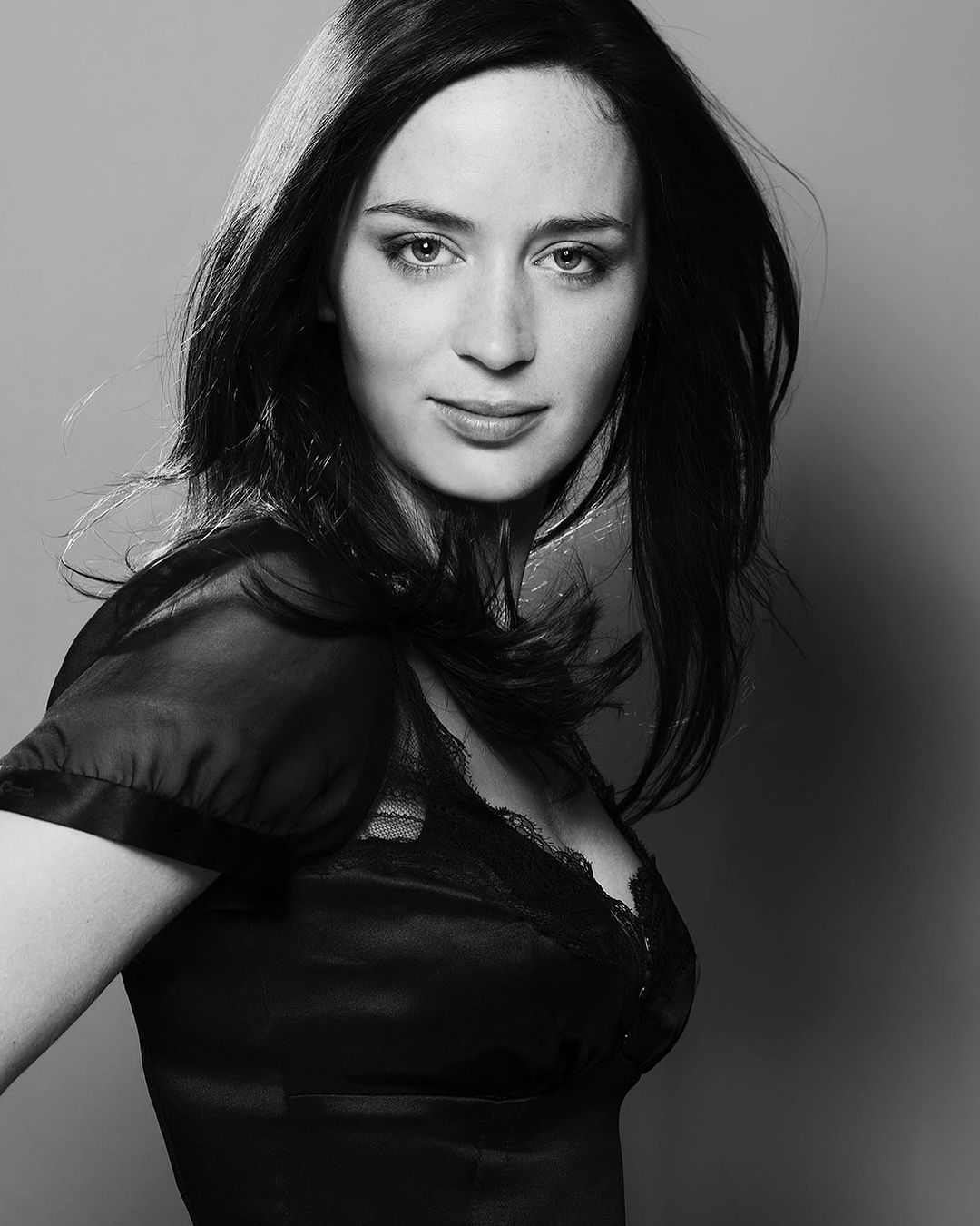 Black and white portrait of an adult female with dark hair, wearing a silk blouse, posing in front of a plain grey background.
