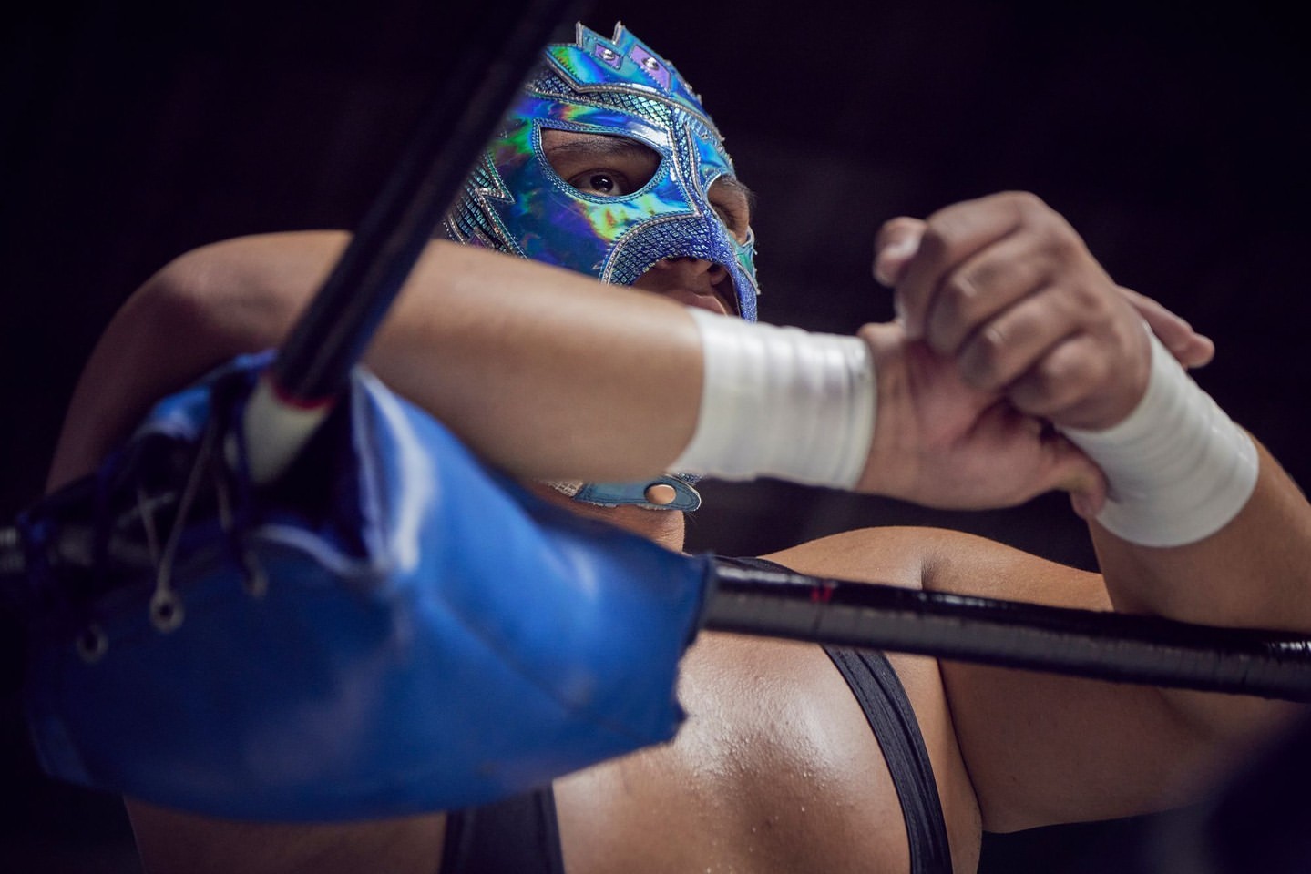 Professional masked wrestler wearing a sparkly blue mask with white wrist wraps in an indoor wrestling environment.