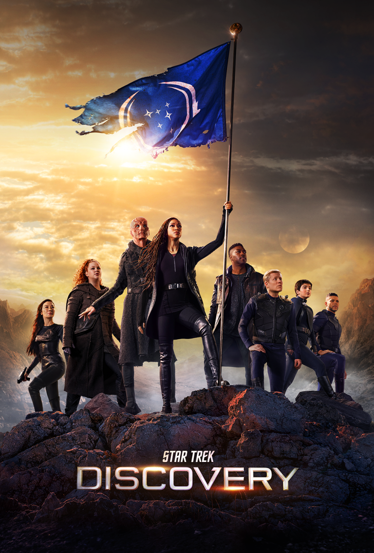 Group of actors posing on a rocky terrain under a blue flag for 'Star Trek Discovery' with a warm sky in the background.