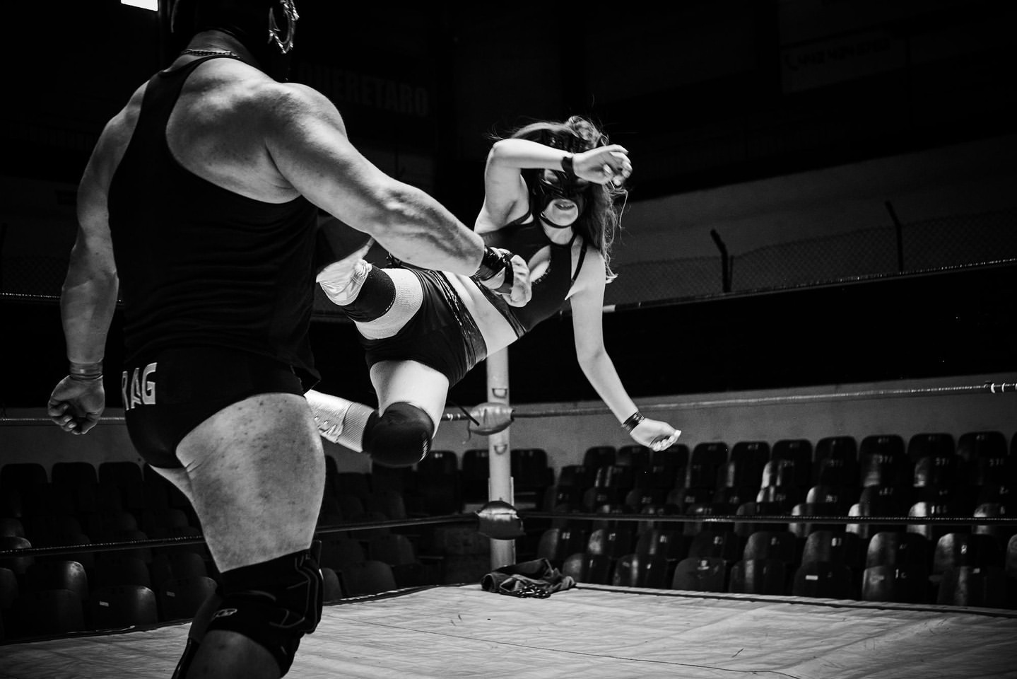 Two wrestlers, one male and one female, are engaged in a wrestling match in a wrestling ring, with the female wrestler kicking.